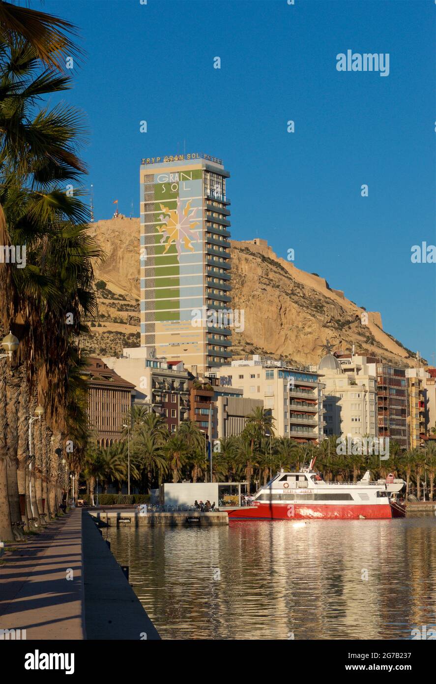 Palm-tree-lined Alicante promenade alongside the marina, Alicante, Valencia, Spain. The 97m high Gran Sol building beyond. Also known as Hotel Gran Sol, Hotel Tryp Gran Sol and officially Edificio Alonso. Mount Benacantil in the background Stock Photo