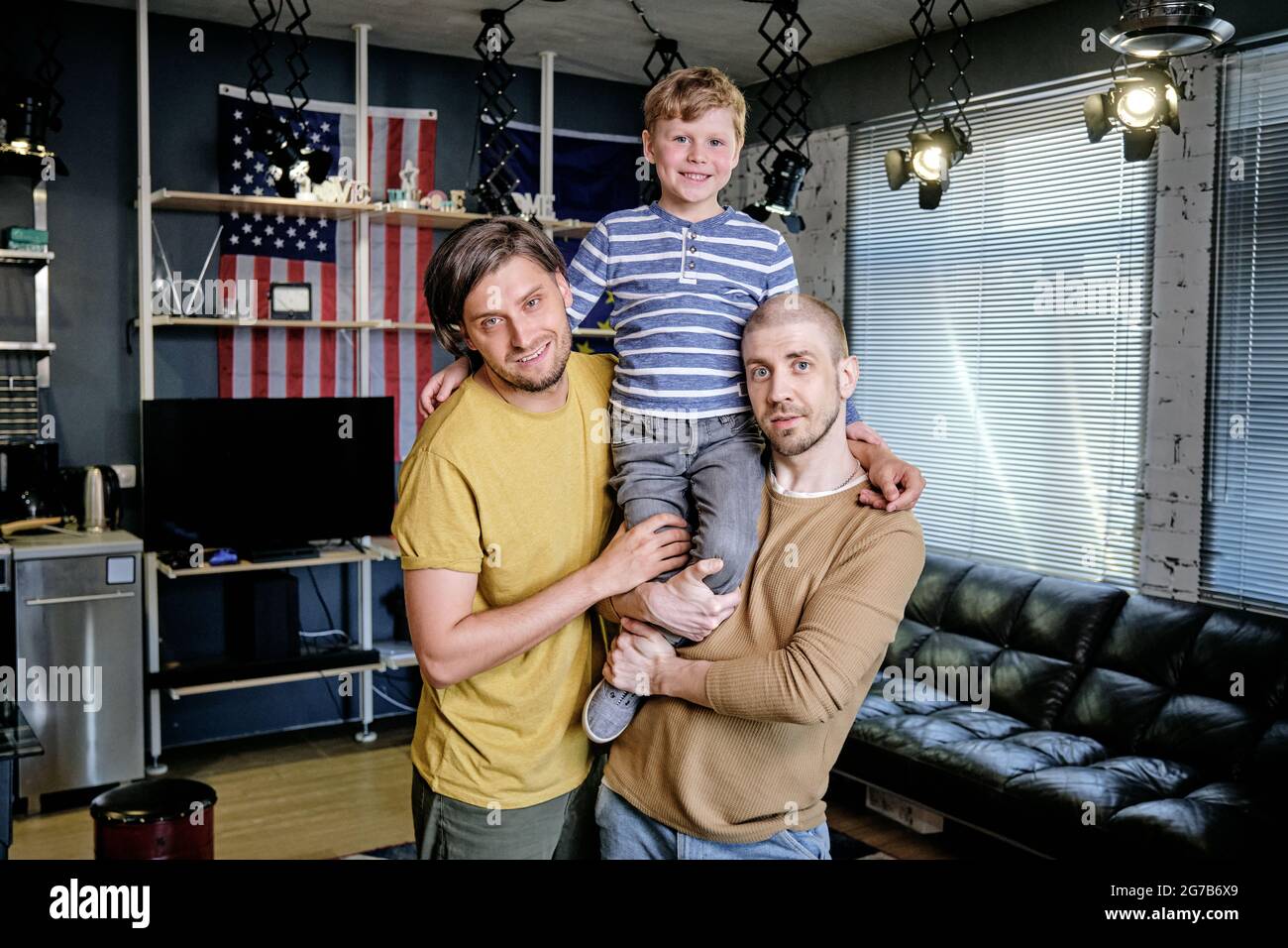Happy young American gay family of three looking at camera while standing in living room of their apartment Stock Photo