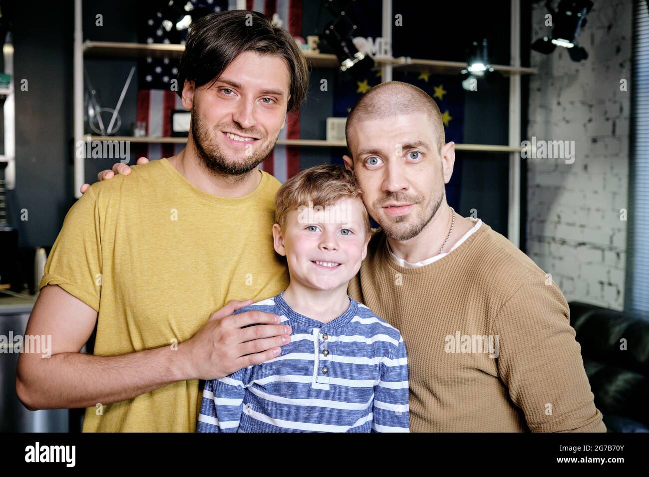 Young gay parents and their little son standing in apartment and smiling at camera Stock Photo