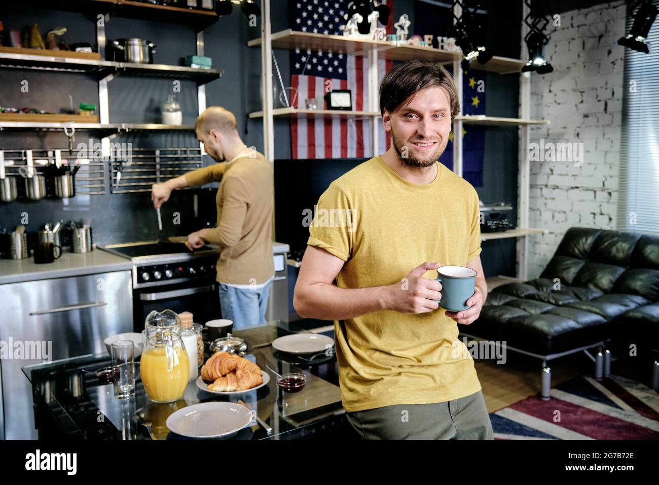 Handsome attractive smiling young man drinking cup morning coffee when his boyfriend cooking breakfast in background Stock Photo