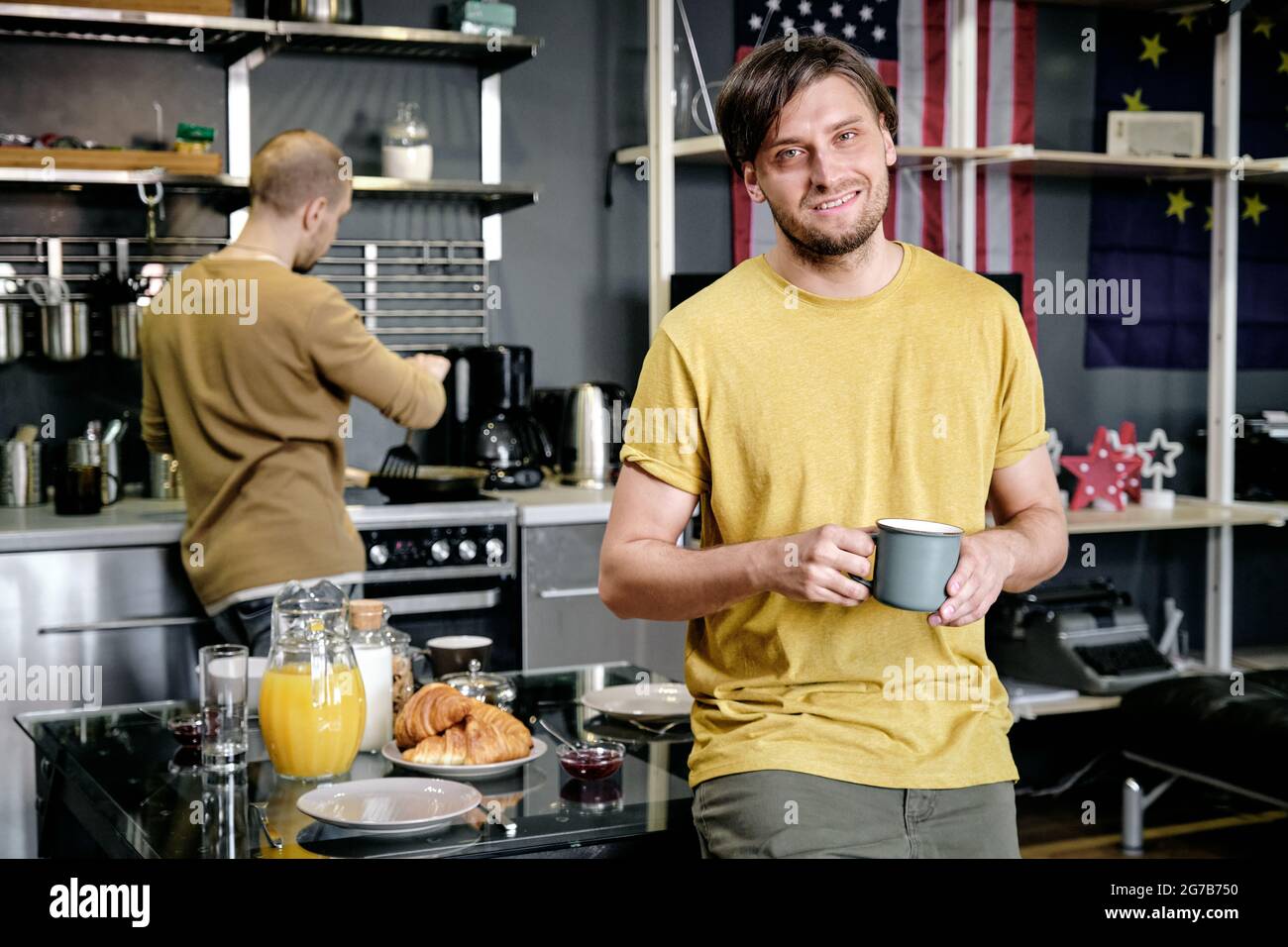 Smiling guy standing in kitchen and having drink, his husband cooking breakfast in background Stock Photo