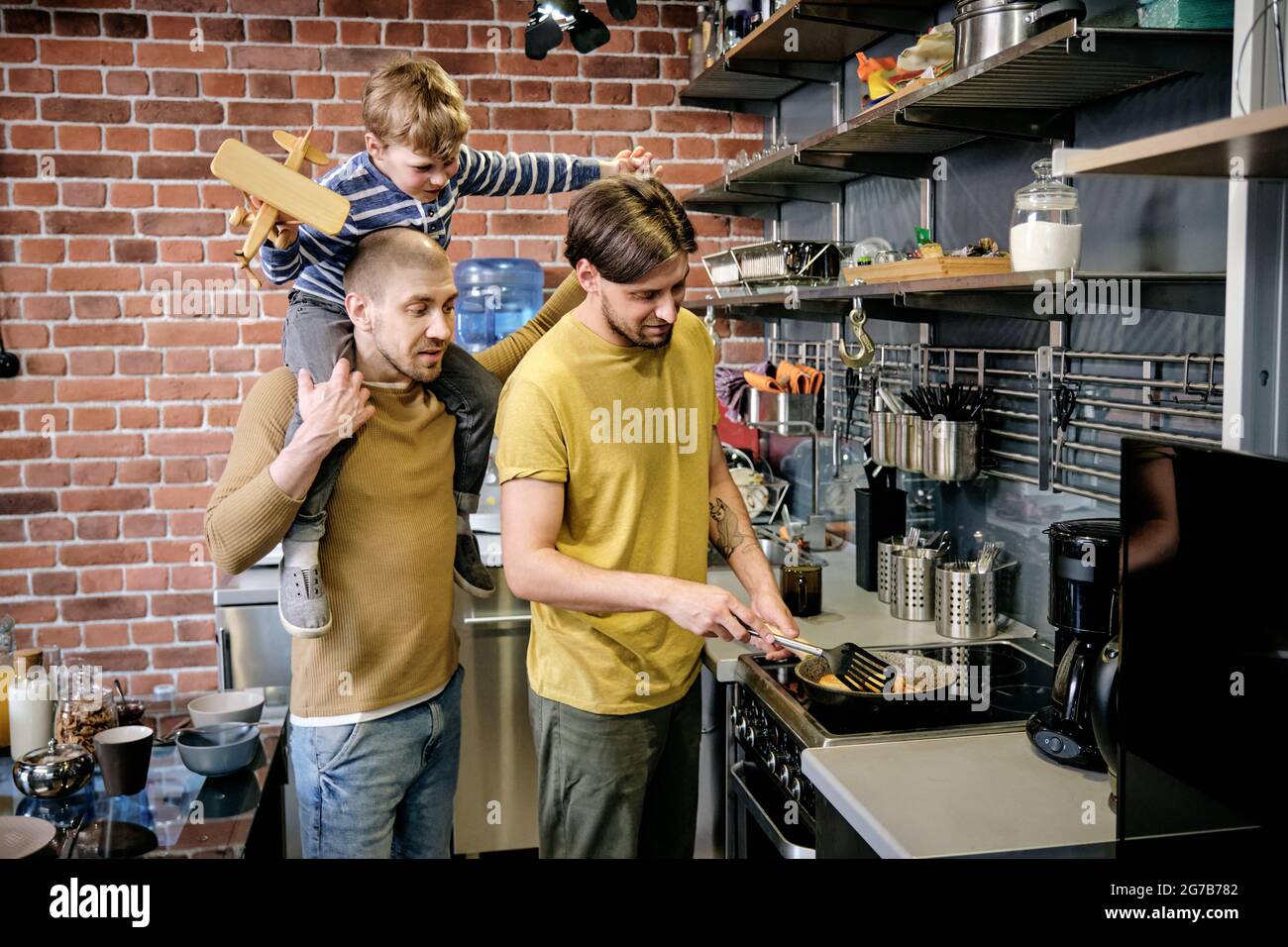 Young gay man cooking breakfast on stove in kitchen when his husband giving piggyback ride to their little son Stock Photo