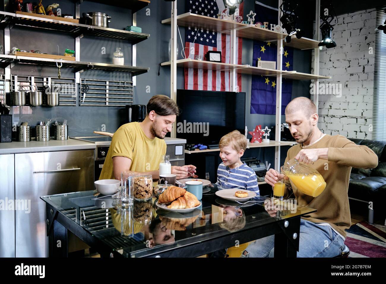 Young man feeding son with breakfast when his husband pouring fresh orange juice in glasses Stock Photo