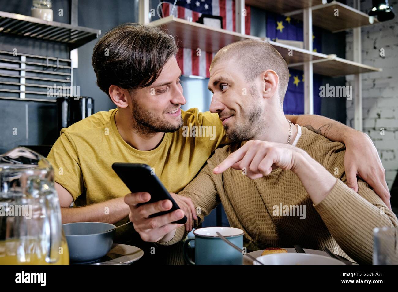Young gay man embracing his boyfriend when discussing new mobile application or news on social media during breakfast Stock Photo