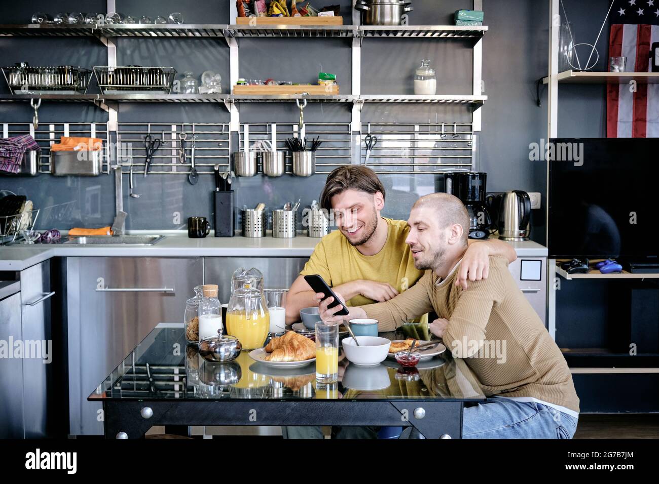 Two young hugging gay men scrolling social media on smartphone when eating breakfast at kitchen table Stock Photo