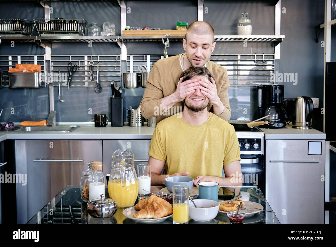 Young man covering eyes of excited boyfriend as he wants to surprise him with tasty homemade breakfast Stock Photo