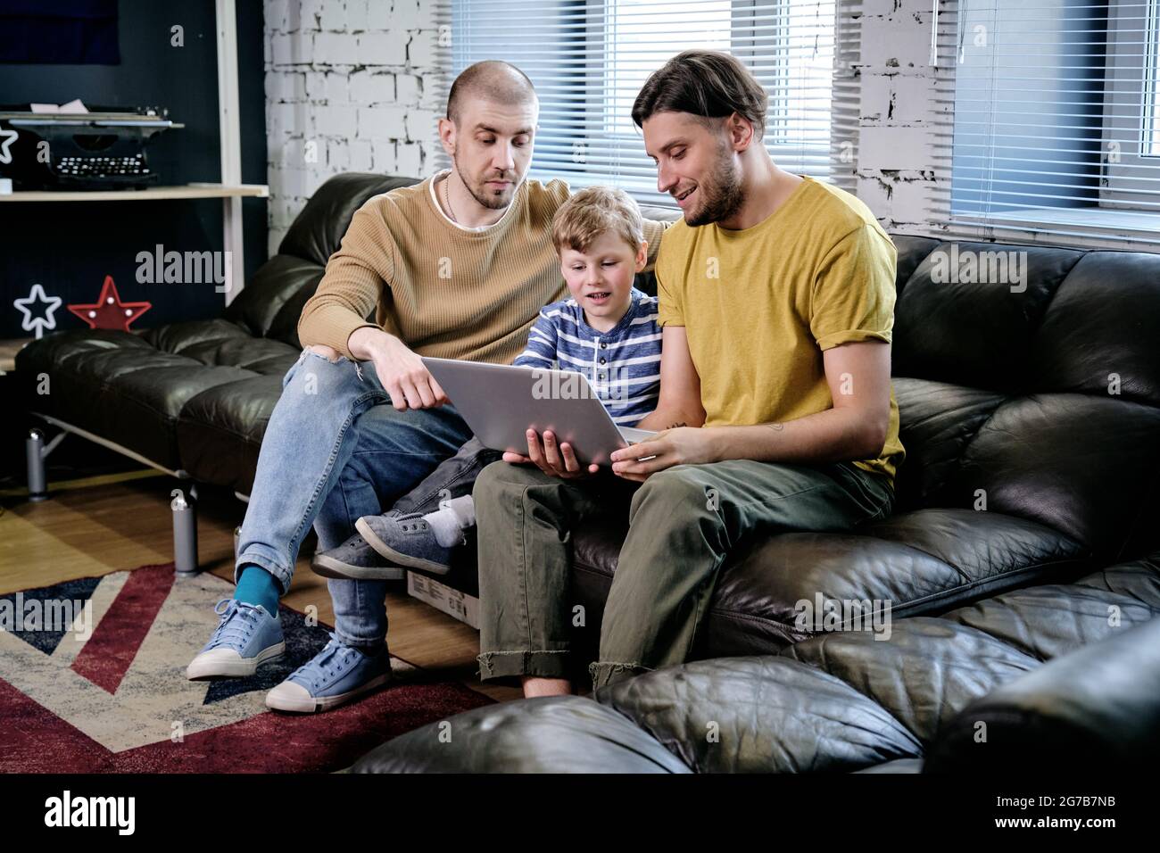 Two young men and little boy sitting on sofa at home and watching animated cartoon or educational video on laptop Stock Photo