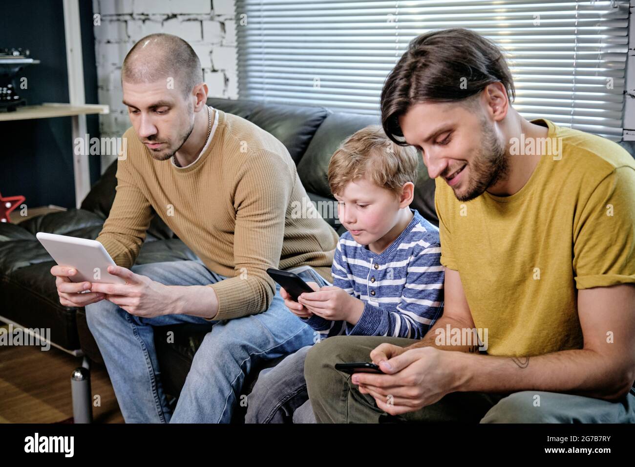 Two young men and little boy using mobile gadgets when sitting on sofa at home Stock Photo