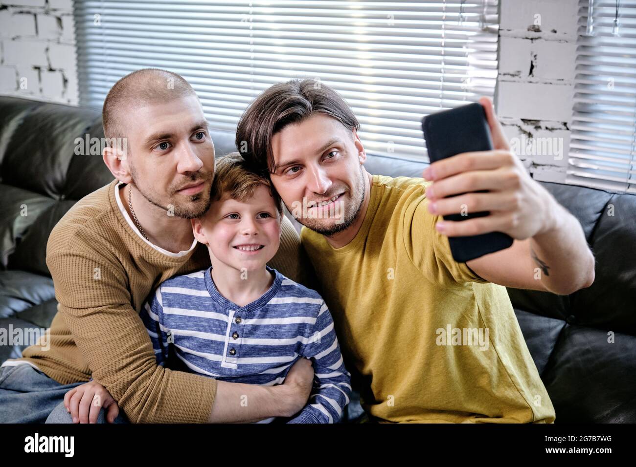 Handsome young man taking selfie with his son and husband when sitting on sofa at home Stock Photo