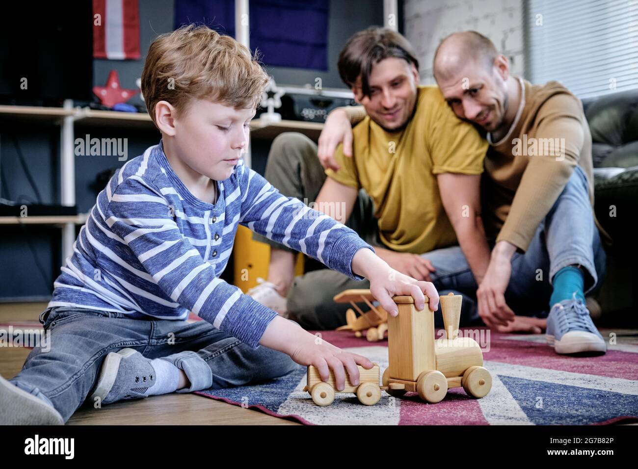 Happy married gay couple looking at their son playing with toys on the floor Stock Photo