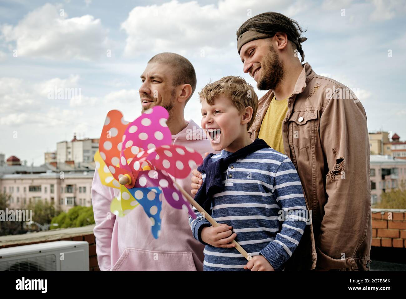 Happy young people and little boy standing on rooftop of building with rainbow pinwheel in hands Stock Photo