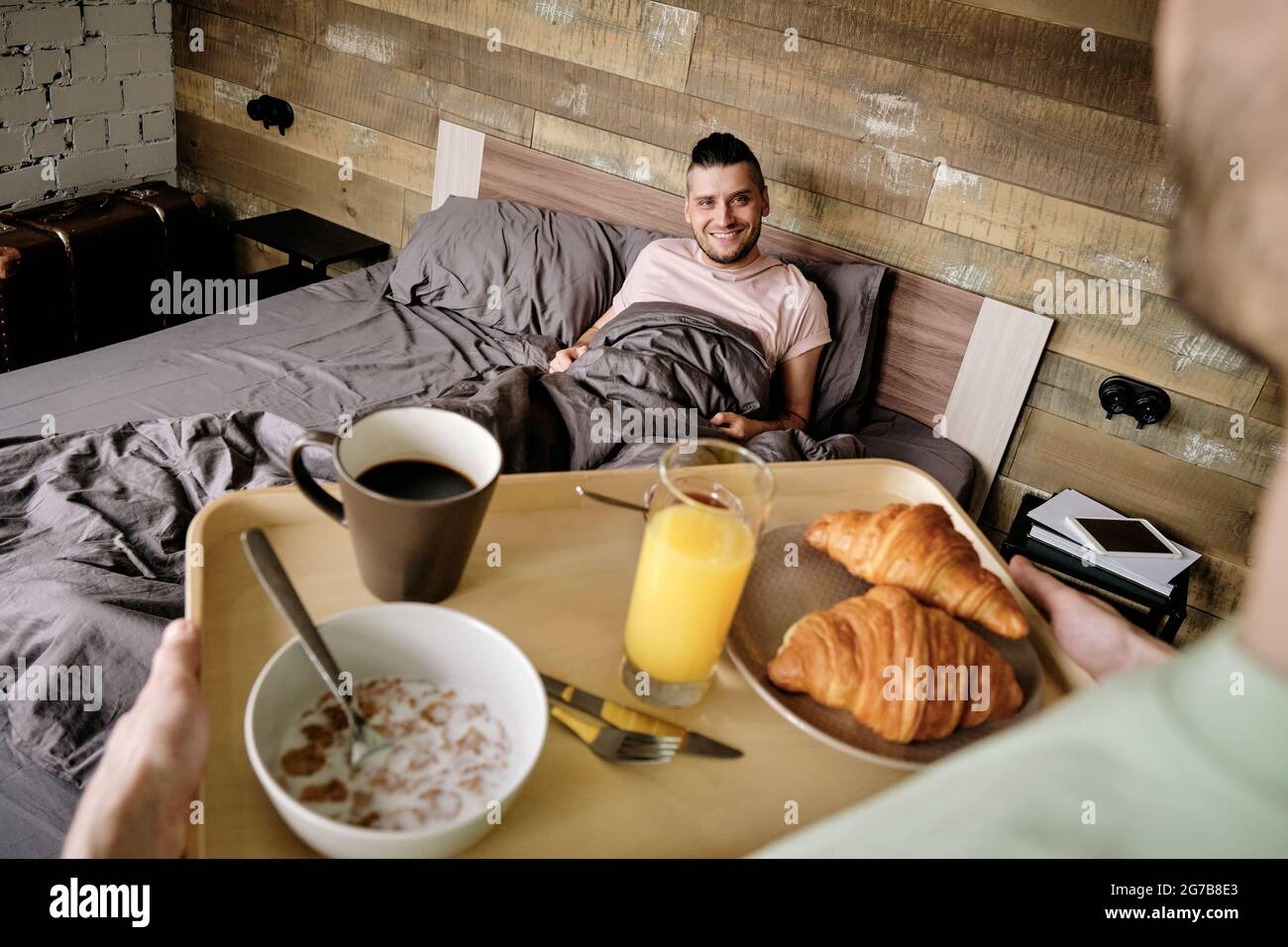 Young man bringing tray with breakfast to his handsome boyfriend in relaxing bed Stock Photo