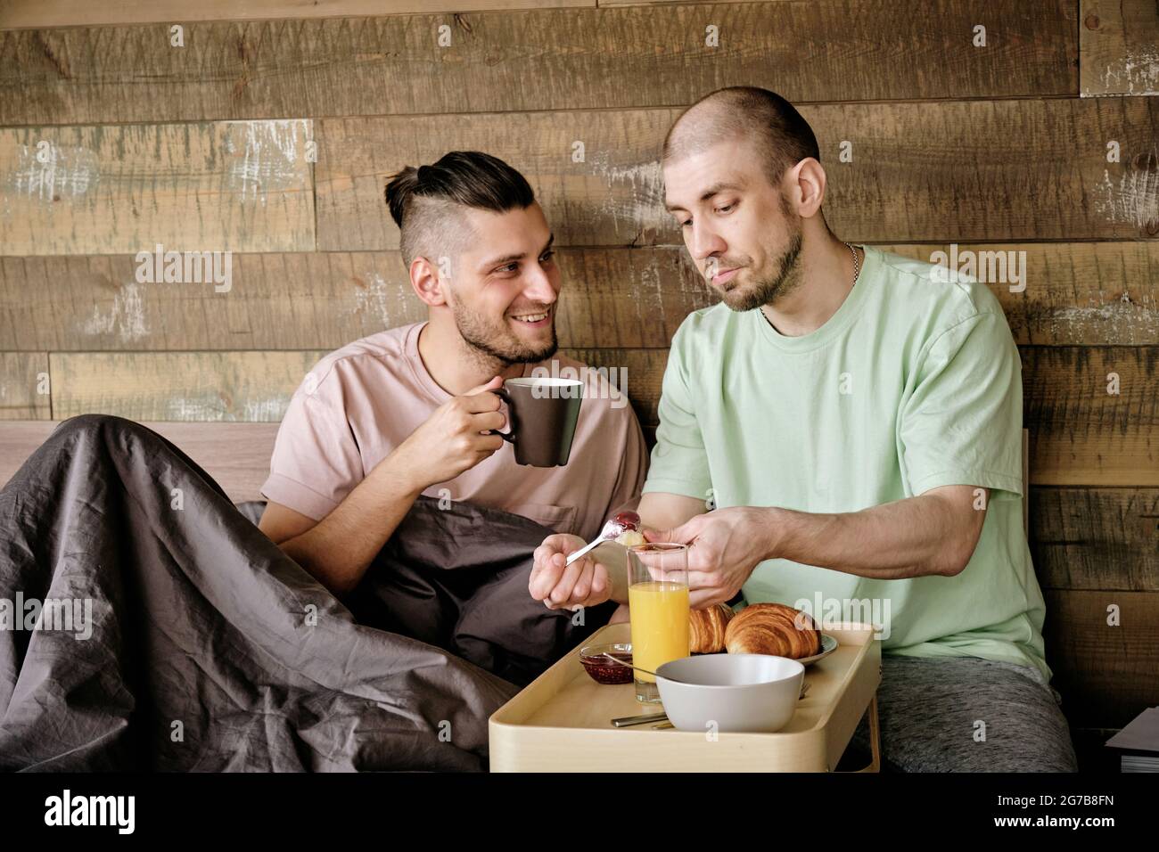 Young gay man putting jam on croissant when his boyfriend drinking cup of coffee and joking Stock Photo