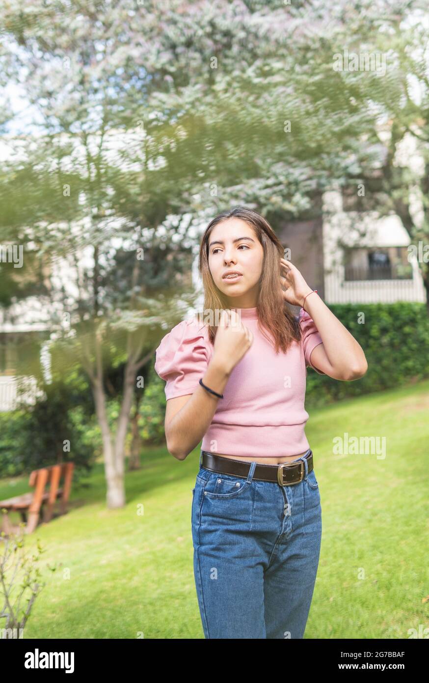 Outdoor portrait of a Latina teen girl with braces adjusting her straight hair with her hands in pink blouse and jeans Stock Photo