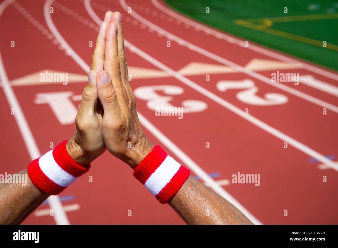 Hands of Japanese athlete wearing red and white colored wristbands making hopeful prayer gesture of gratitude against a red athletic track background Stock Photo