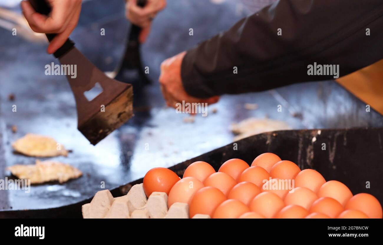 A tray or carton of eggs in the foreground with a hot plate bbq in the background. hands and cooking tools turning fried eggs. Community fundraiser Stock Photo