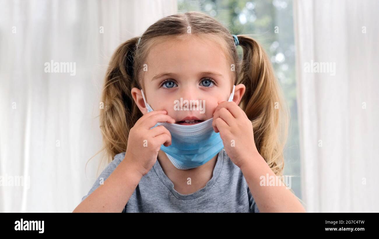 Little cute girl takes off her medical mask, smiles and puts it back on her face with ponytails. Child is sick at home Stock Photo