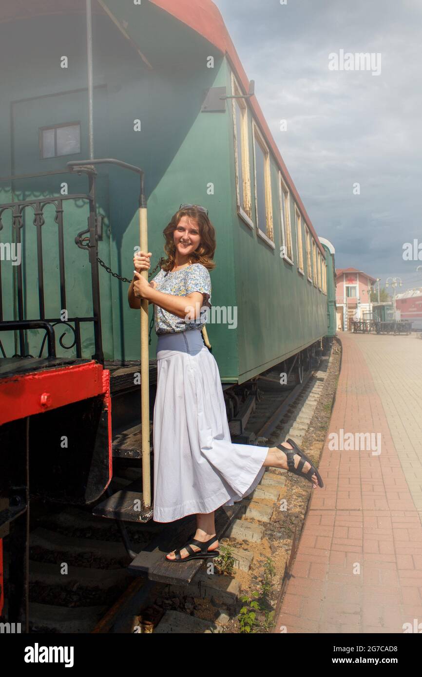 Woman standing on the running Board of the car. Holding the handrail of the departing train. Empty platform in vintage style. Retro cars and locomotiv Stock Photo