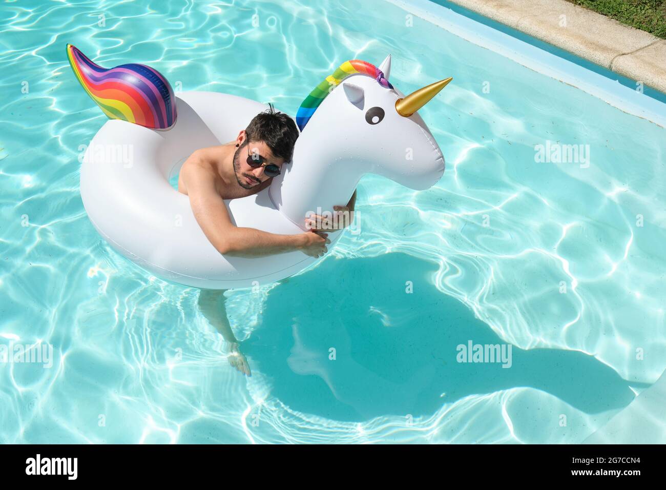 Young man wearing sunglasses hugging a big unicorn inflatable ring in a swimming pool. Summer concept. Stock Photo