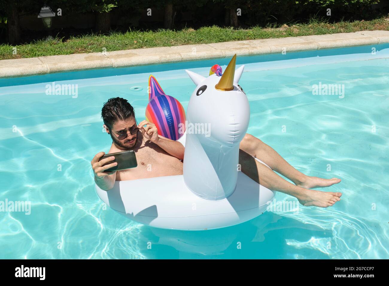 Young man wearing sunglasses taking a selfie on a unicorn inflatable ring in a swimming pool. Summer concept. Stock Photo