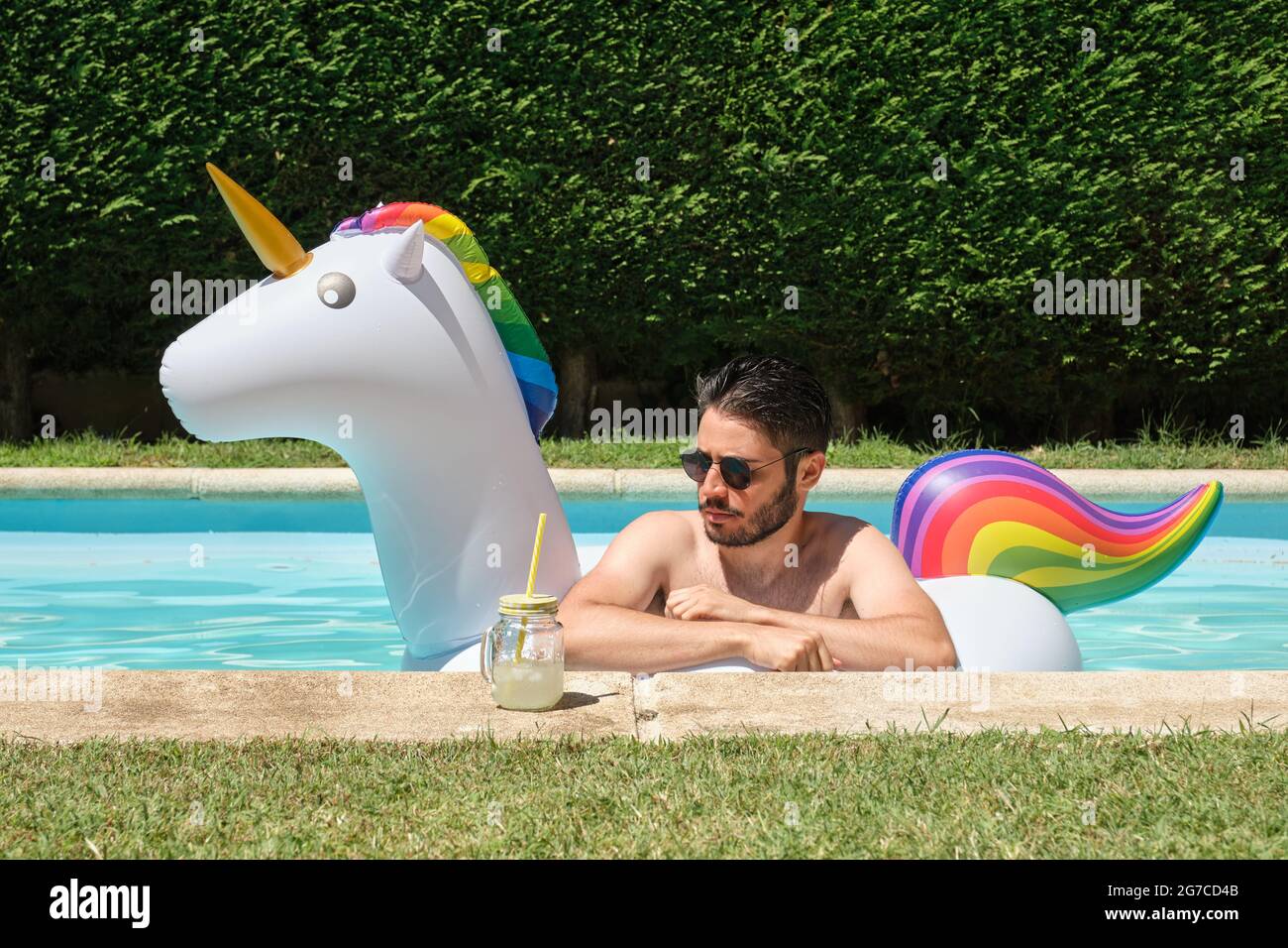 Young man wearing sunglasses into a unicorn inflatable ring in a swimming pool with a refreshment. Summer concept. Stock Photo