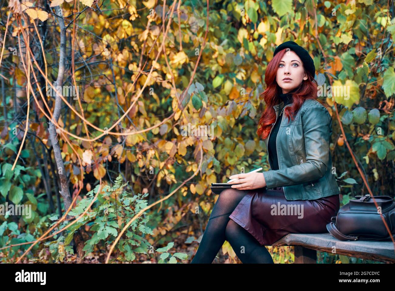 Woman with graphic tablet on park bench. Autumn landscape in the background. Red-haired girl in a beret and a fashionable jacket with a leather backpack. Designer or freelancer in nature. Stock Photo