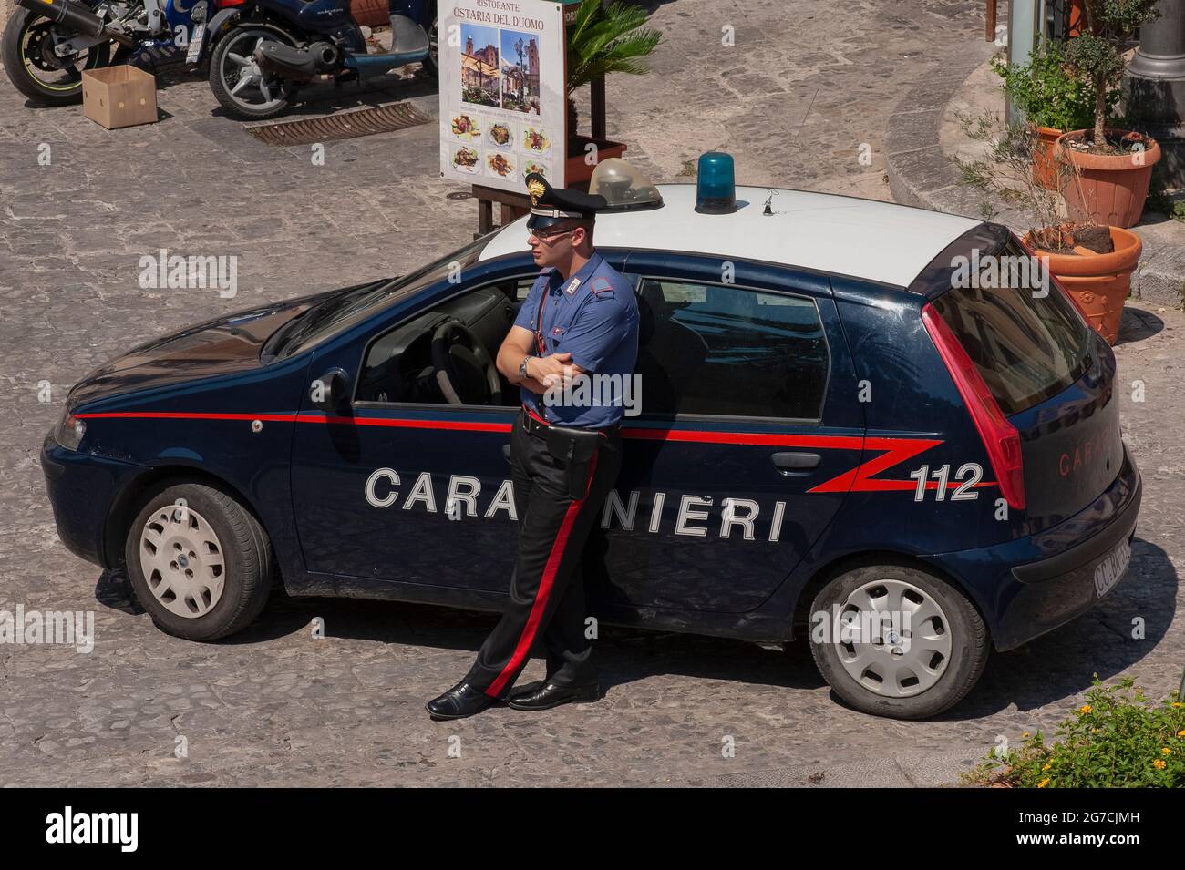 A young Carabiniere, an armed officer in Italy's Arma dei carabinieri military  police force, leans against his Fiat patrol car with arms folded as he  keeps a watchful eye on tourists walking