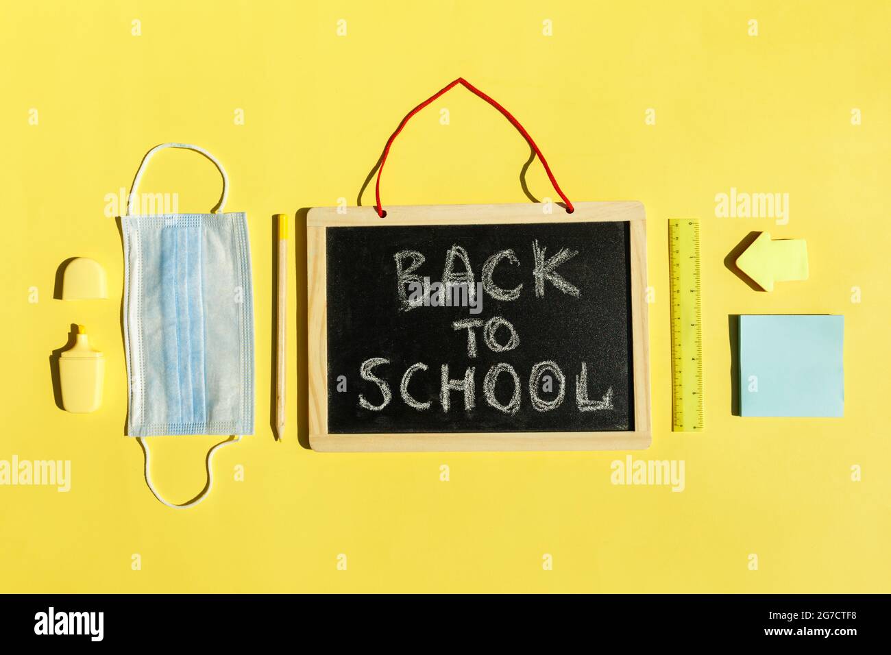 School stationery supplies, medical mask on yellow background social distancing, school reopening after covid-19 pandemic. New normal concept.Top view copy space,blackboard. Stock Photo