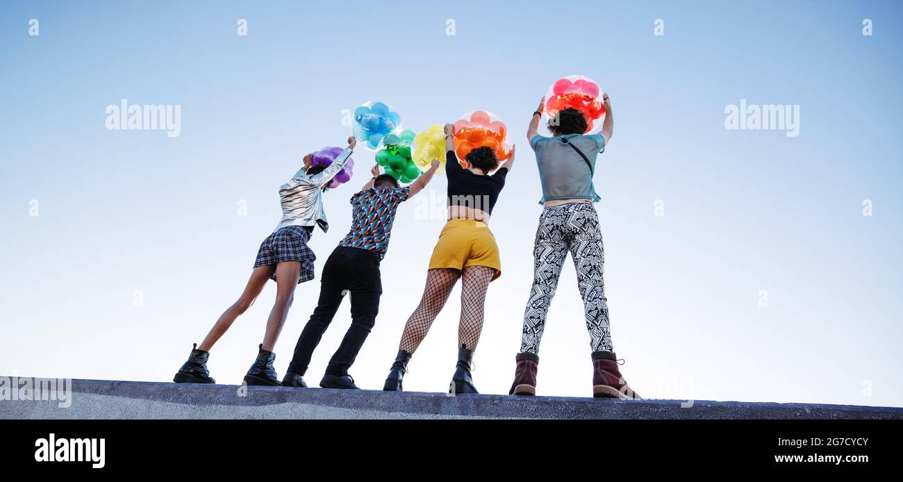 Unrecognizable queer people embracing gay pride. Group of four friends raising up balloons with rainbow colours outdoors. Young members of the LGBTQ+ Stock Photo