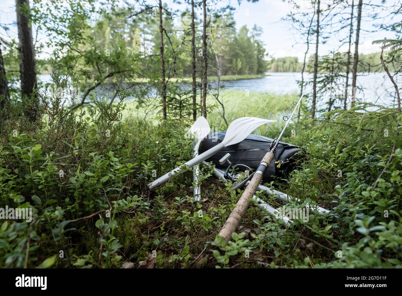New rolled up rubber inflatable boat, oars and a spinning rod lie in the grass, on the shore of the lake. Preparing for fishing. Stock Photo