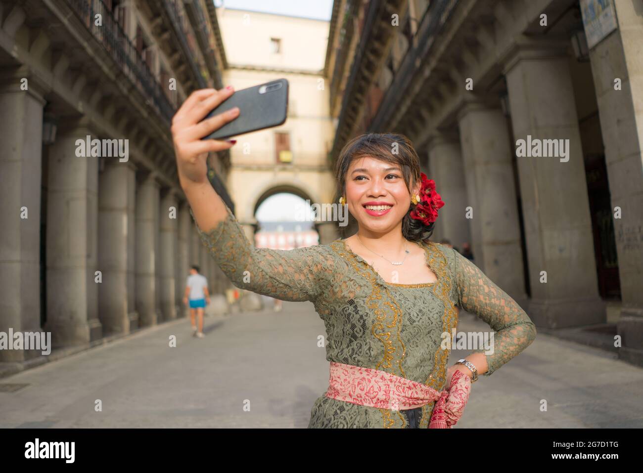 young happy and beautiful Asian woman wearing traditional Balinese kebaya dress taking selfie - Indonesian girl in Bali clothes walking on city street Stock Photo
