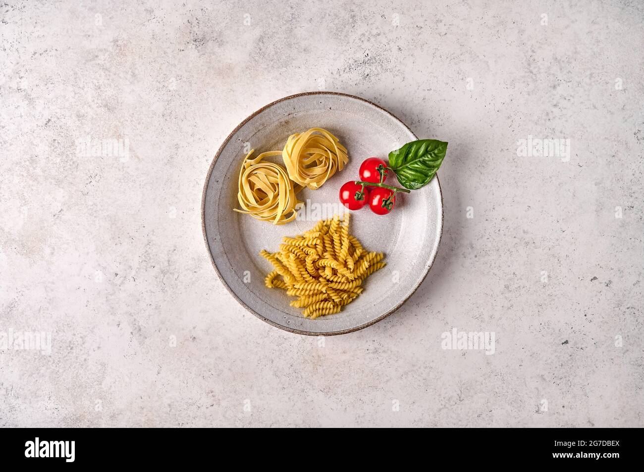 Uncooked raw girandole, tagliatelle, cherry tomatoes and basil on white plate on grey textured background. Copy space. Top view Stock Photo