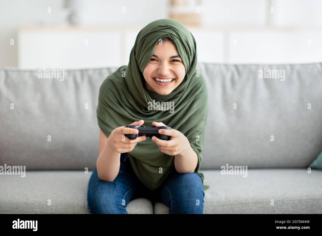 Portrait of happy teen girl in hijab holding joystick, playing videogame at home. Lockdown pastimes and hobbies Stock Photo