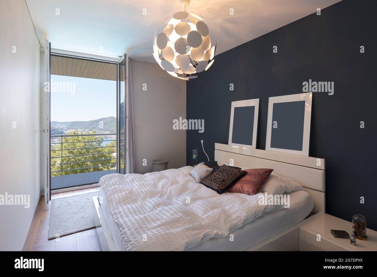 Elegant bedroom with large white bed and a balcony with a view of the valley with the lake. Nobody inside. Stock Photo