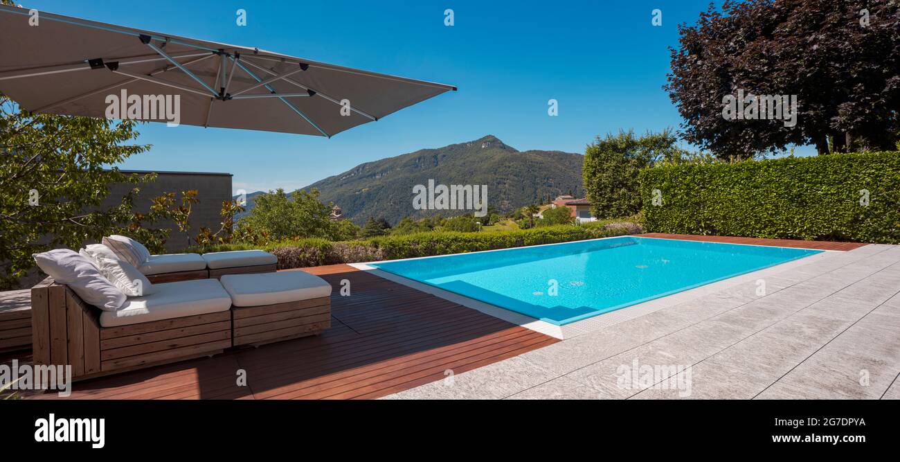 Large modern pool with two sun loungers for sunbathing and an open umbrella. View of the mountains of Switzerland. Nobody inside. Stock Photo