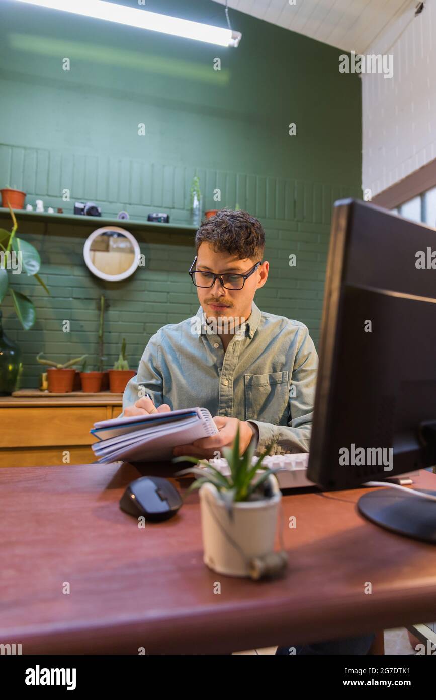 Young Hispanic male writing a note in the notepad sitting in front of ...