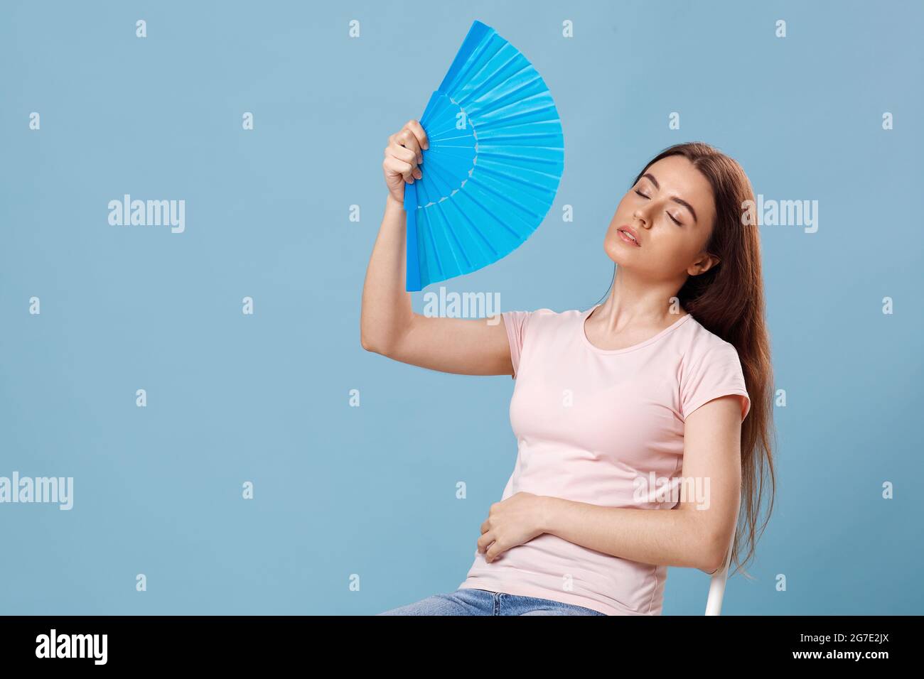 Overheated young lady sitting on chair with paper fan Stock Photo