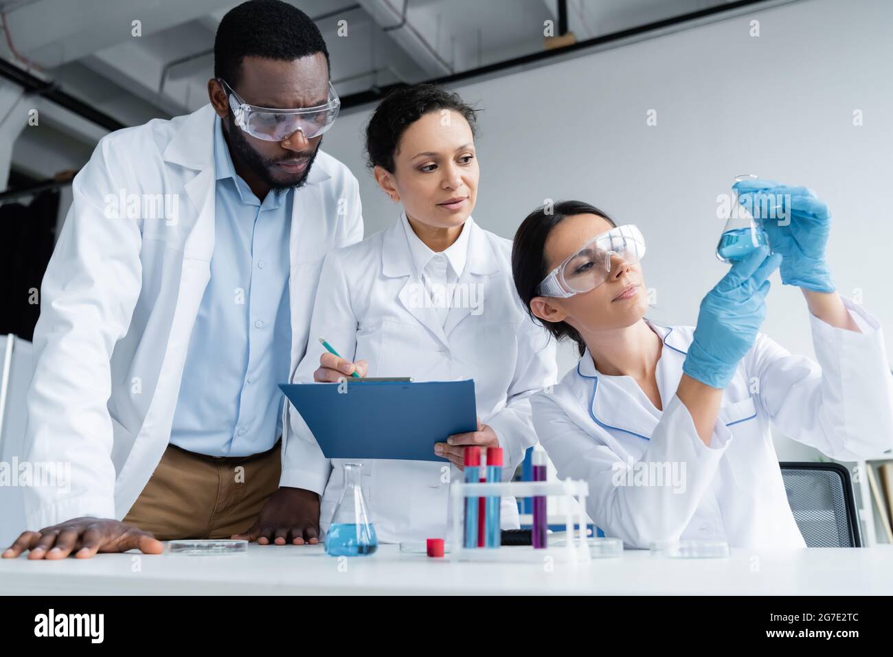 Interracial scientists in safety goggles with clipboard looking at reagent in flask Stock Photo