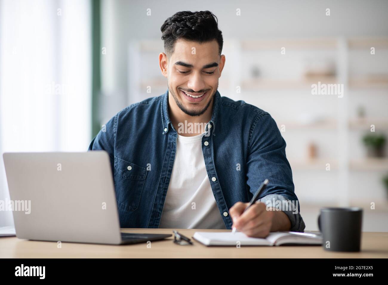 Joyful arab man taking notes while working with laptop Stock Photo