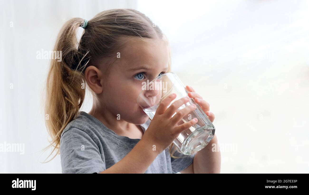 Little blonde girl in white dress drinks water from a glass indoors. Cute child is drinking a cup of water Stock Photo