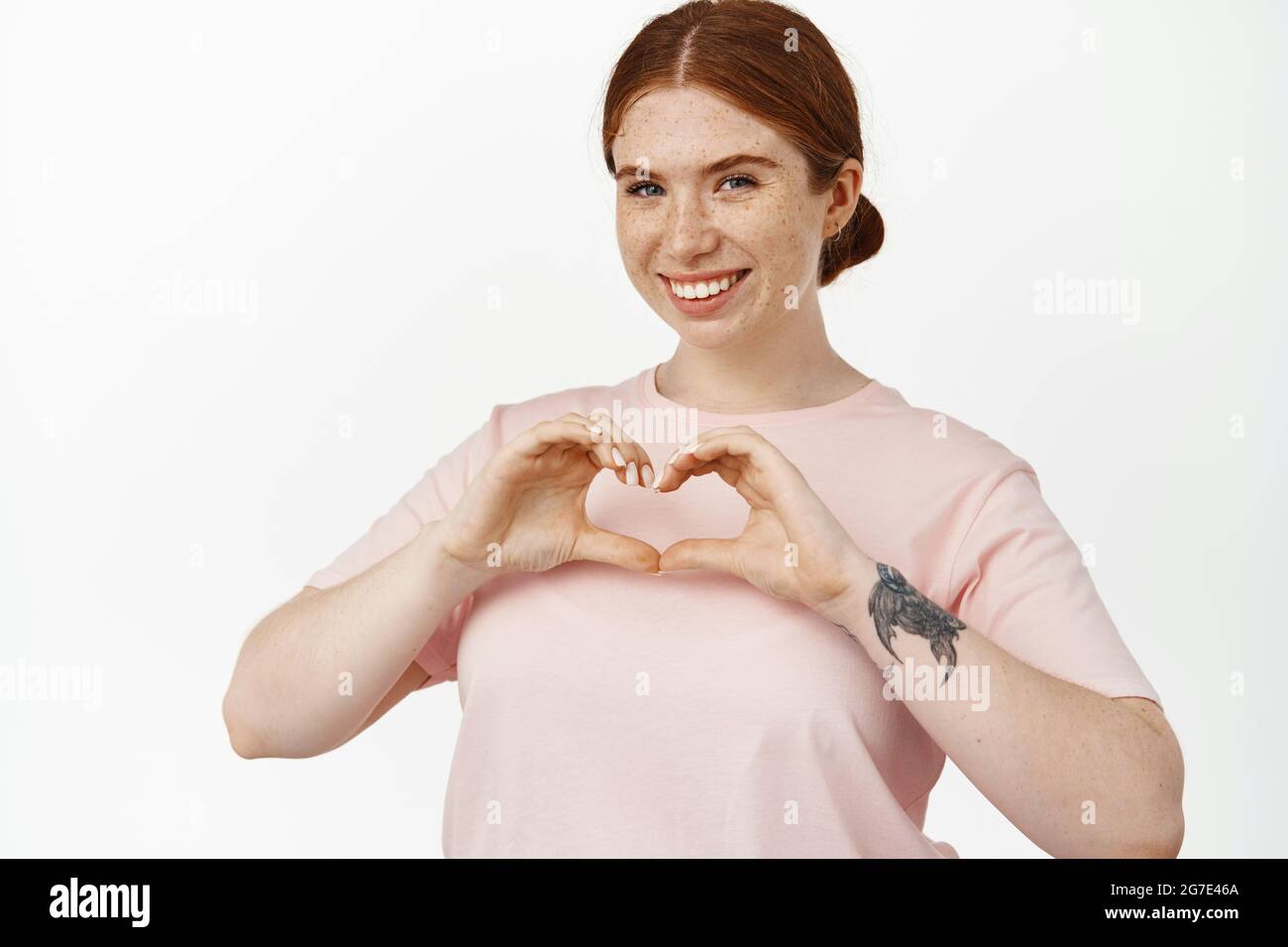 In love. Young smiling redhead female shows heart sign and looks happy, like something, express affection or sympathy, standing in tshirt against Stock Photo