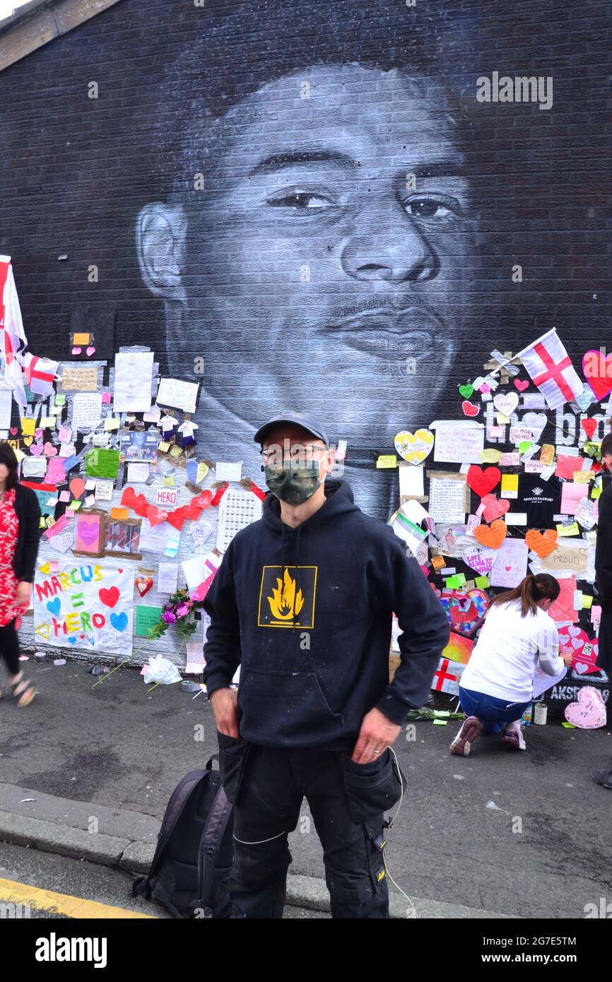 Street artist Akse P19 in front of the giant Manchester United player  Marcus Rashford mural in Withington, Manchester, England, United Kingdom, which he painted. The mural was vandalised  with abusive graffiti after England's Euro2020 football loss on July 11th, 2021. The mural was created by French-born street artist Akse on the wall of the Coffee House Cafe on Copson Street. Marcus Rashford is a Manchester United football player. Akse fully repaired the damaged mural by 13th July, 2021. Picture date: July 13th, 2021. Stock Photo