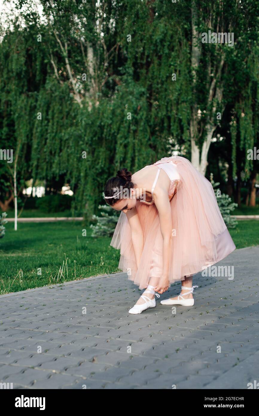 lifestyle full-length portrait of a ballerina in a pink dress with a full organza skirt bends down to her leg to tie ribbons on pointe shoes or ballet Stock Photo