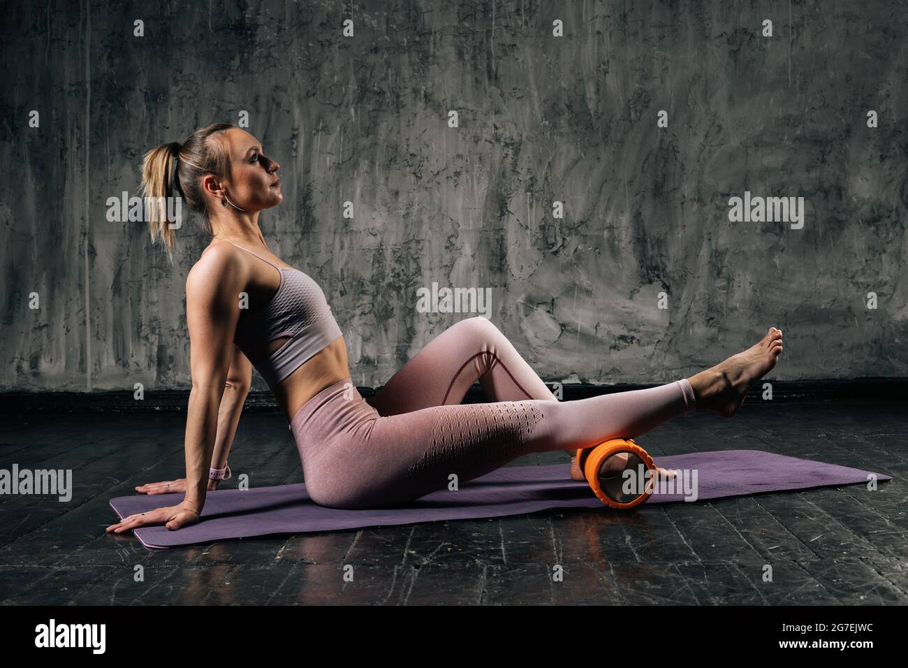 Muscular young athletic woman with perfect beautiful body wearing sportswear using foam roller massager sitting on yoga mat. Stock Photo