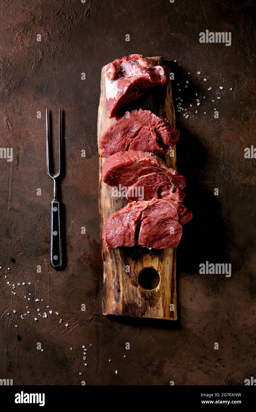 sliced raw beef tenderloin meat for steaks on wooden board with metal meat fork, salt and pepper over dark brown texture background. Food cooking back Stock Photo
