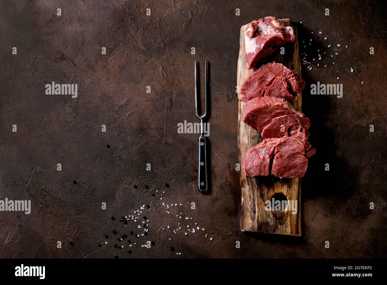 Fresh sliced raw beef tenderloin meat for steaks on wooden board with metal meat fork, salt, pepper over dark brown texture background. Food cooking b Stock Photo