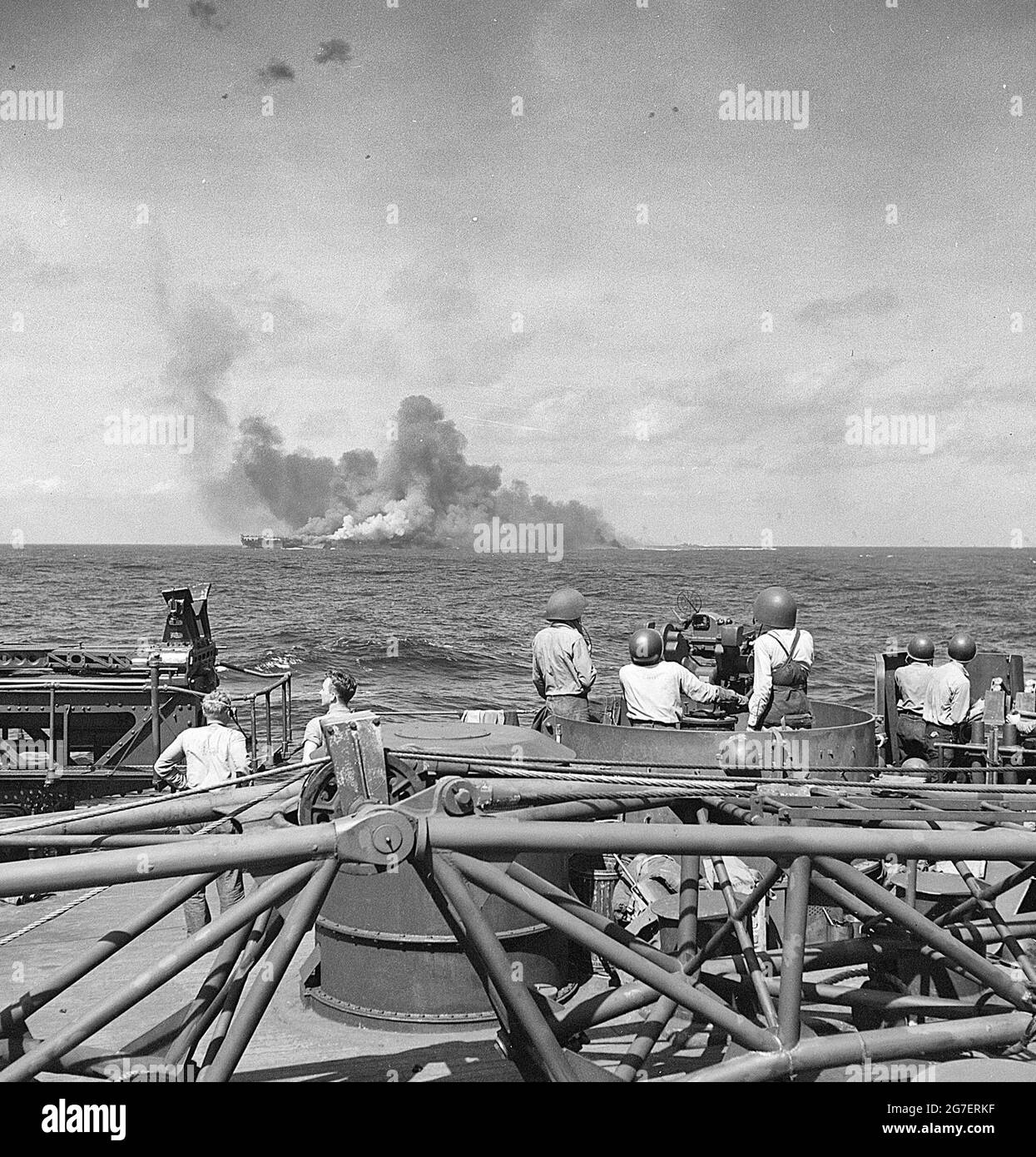 USS Intrepid (CV-11) after being hit by Japanese plane in suicide dive in the Pacific. Taken from USS New Jersey (BB-62). Smoke coming from USS Intrepid (CV-11) gunners at 40mm guns on board USS New Jersey (BB-62) in foreground Stock Photo
