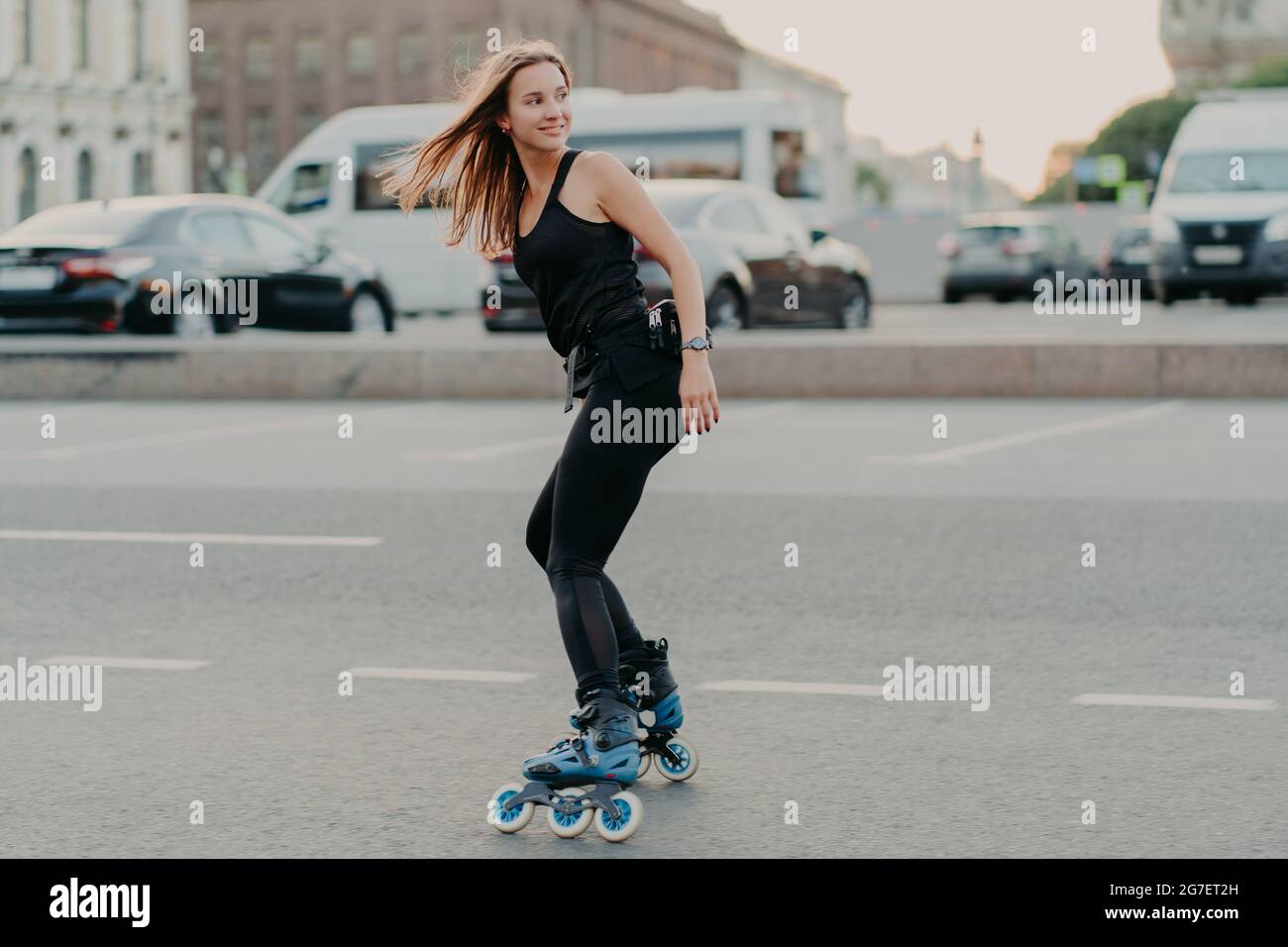 Sideways shot of active young woman in good physical shape rollerblades along city streets looks happily behind wears black sportsclothes leads Stock Photo