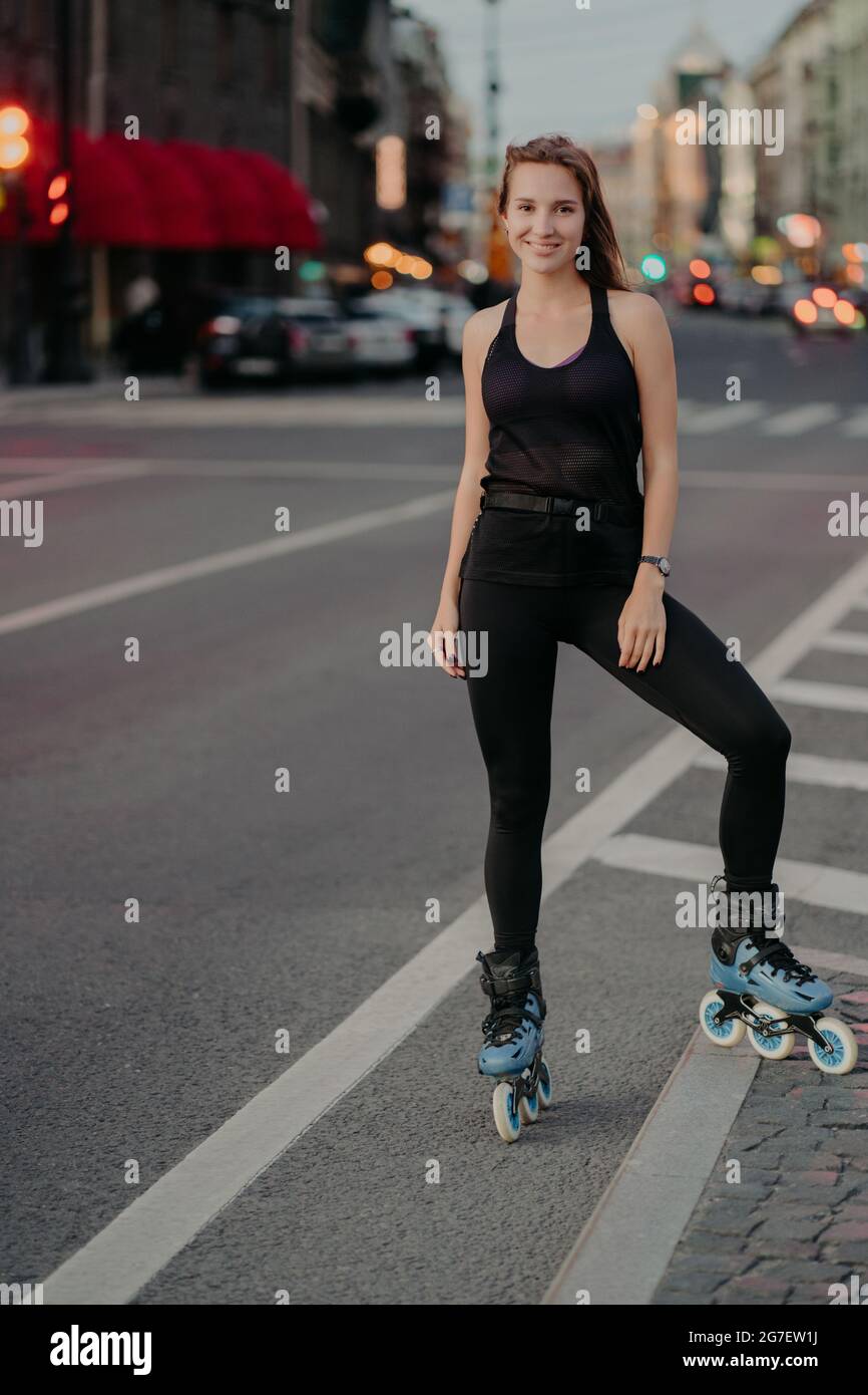 Full length shot of active young woman being in good physical shape dressed in black activewear enjoys rollerblading during good summer day poses on Stock Photo