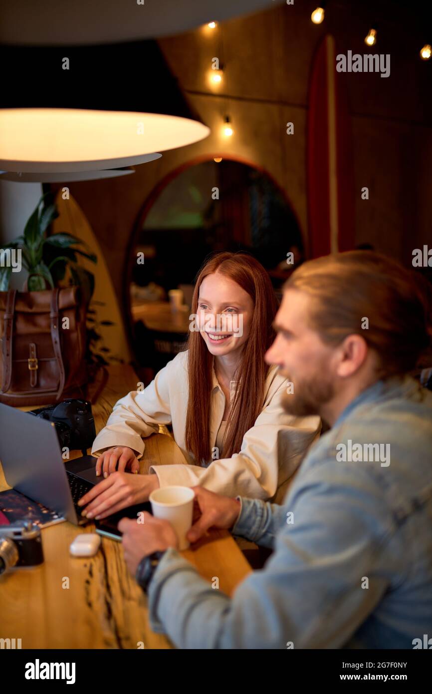 Side view portrait of cheerful caucasian managers photographers redhead female and bearded guy working in cafe, use laptop, surfing internet, having t Stock Photo